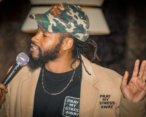 Ken Jones performing live with a microphone, wearing a camo “Pray My Stress Away” hat and tan coat, mid-speech with expressive hand gesture.